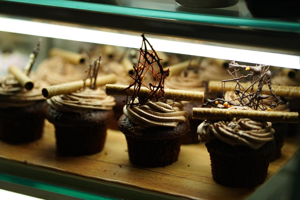 Šokolādes kēksi ar radošām dekorācijām, tostarp šokolādes stieņiem un saldām spirālēm. Kēksi ir izkārtoti uz koka plaukta, radot pievilcīgu izskatu.