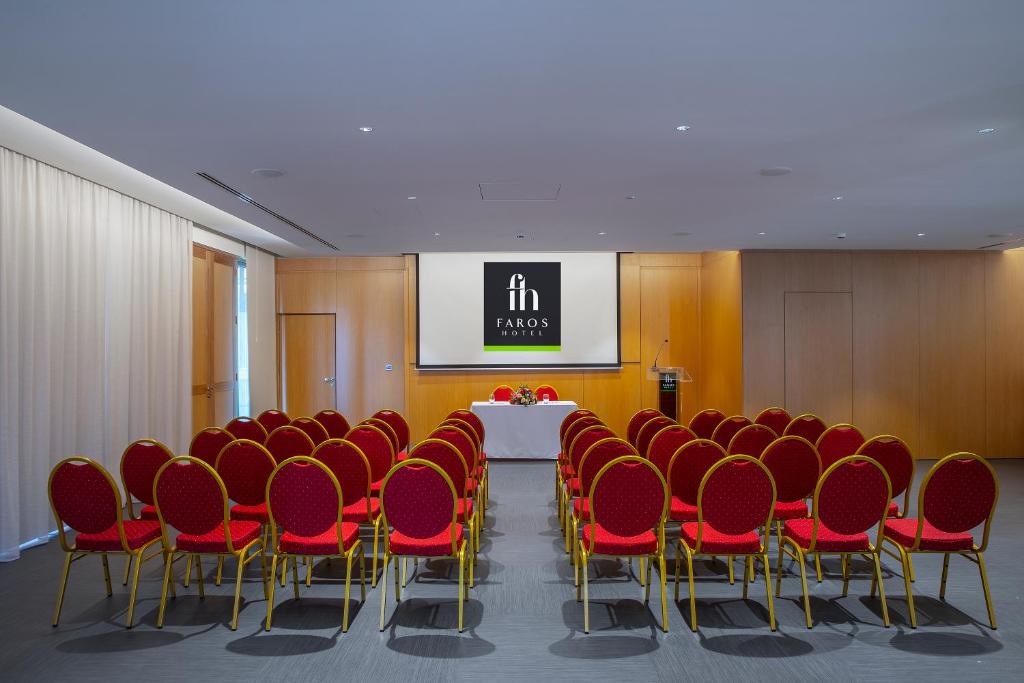 Konferenču zāle ar sarkaniem krēsliem un ekrānu, uz kura redzams "Faros Hotel" logo. Zāle ir sagatavota pasākumam ar galdu priekšā.