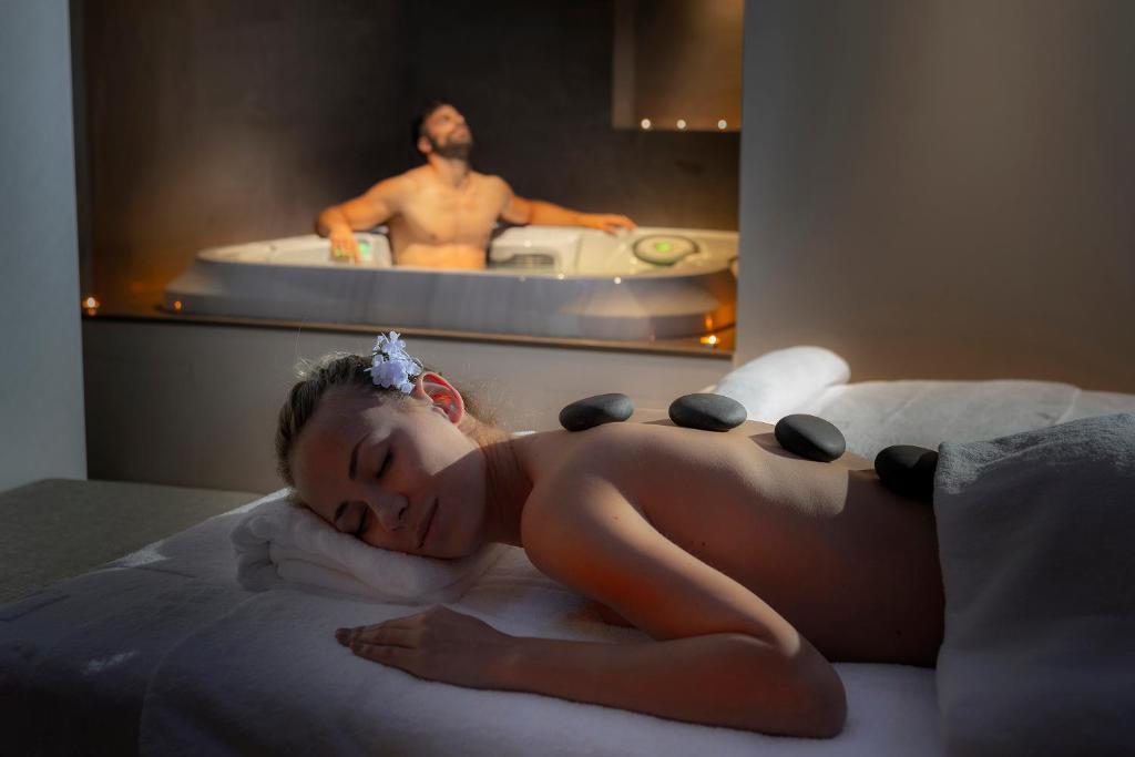 Mierinoša spa atmosfēra ar relaksējošu masāžu, kur sieviete guļ uz masāžas galda ar akmeņiem uz muguras, bet vīrietis atpūšas džakuzi.