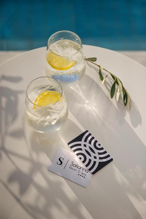 Divi dzērieni ar citrona šķēlēm un ledus gabaliņiem uz galda, blakus atpūtas kartēm no Solanna Resort & Spa. Fons ar baseinu papildina relaksējošu atmosfēru.