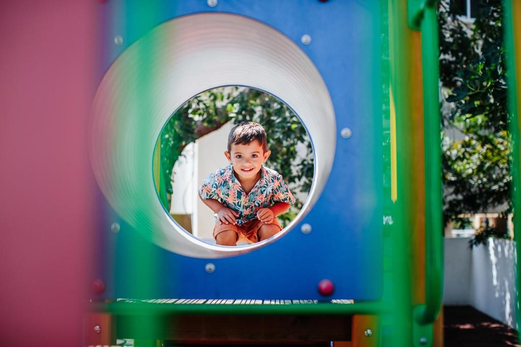 Bērns rotaļu laukumā, sēžot caurspīdīgā caurumā, ar priecīgu smaidu un krāsainu kreklu. Rotaļu aprīkojums ir krāsains un apkārt ir zaļumi.