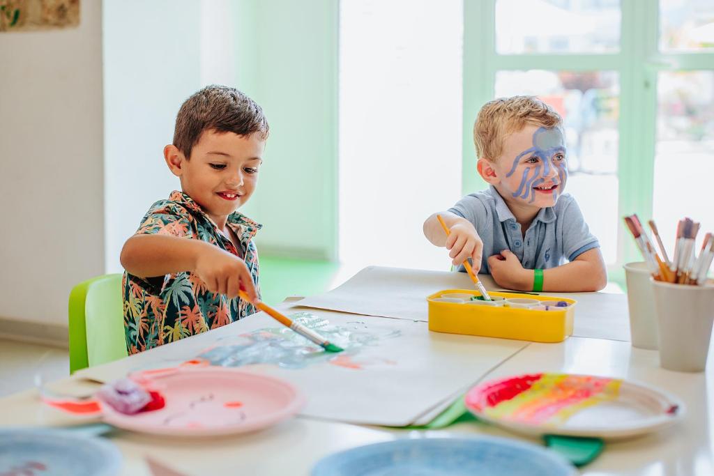 Divi bērni radoši glezno, izmantojot krāsas un otas, priecājoties un smējoties. Viens no bērniem ir ar krāsainu sejas zīmējumu.