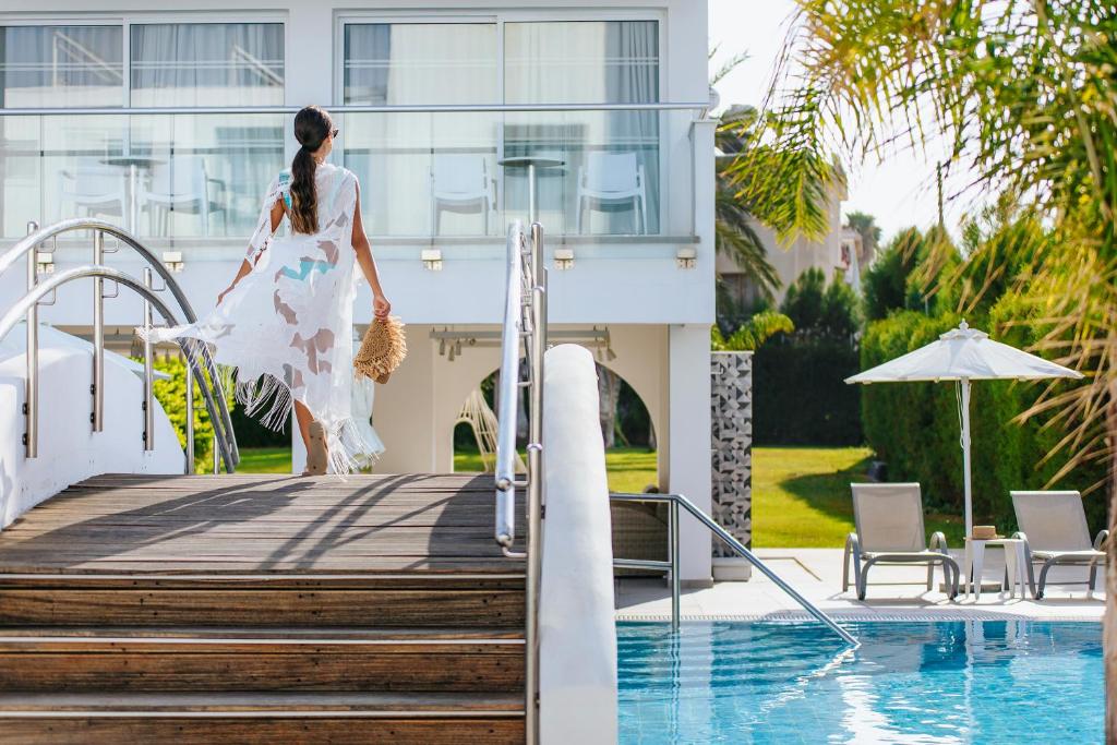 Sieviete ar plūstošu apģērbu dodas uz baseinu, baudot saulainu dienu elegantā un modernā vidē. Apkārt ir zaļi zālieni un baseins, kas rada atpūtas atmosfēru.