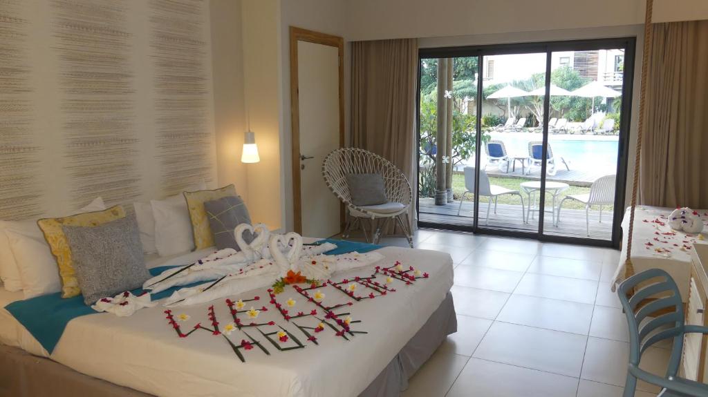 Romantiski iekārtota viesnīcas numuriņš ar gultu, dekorētu ar ziediem un dvieļu sirds formām, un skatu uz baseinu. Interjers ir gaišs un mūsdienīgs, radot relaksējošu atmosfēru.