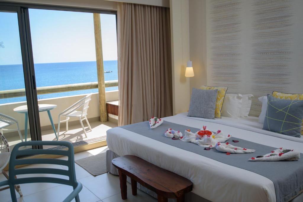 Mūsdienīga viesnīcas numura interjers ar skatu uz jūru, kas iekārtots ar ziedu dekorācijām uz gultas. Logi ved uz balkonu ar ērtām mēbelēm un saulainu skatu.