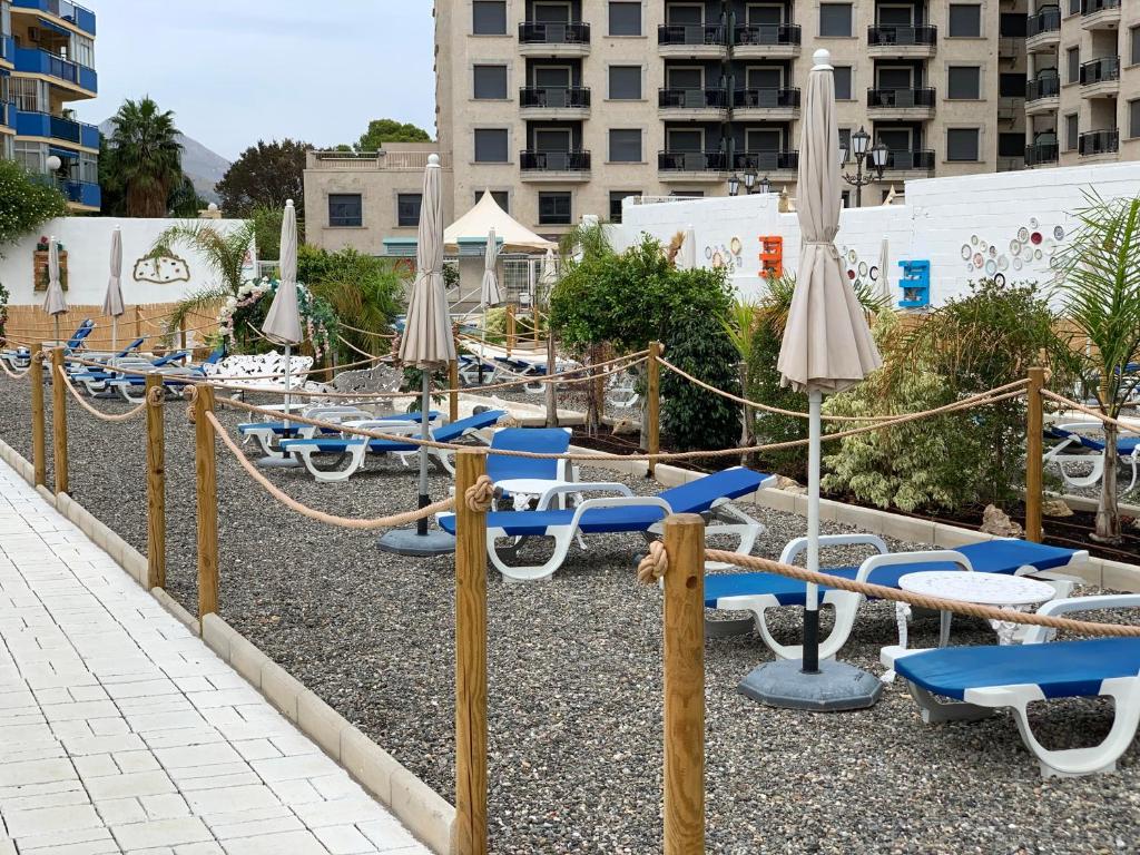 Atpūtas zona ar sauļošanās krēsliem, lietussargiem un zaļiem augiem, kas izveidota pie viesnīcas. Apkārt ir akmeņi un koka žogs, radot mierīgu atmosfēru.