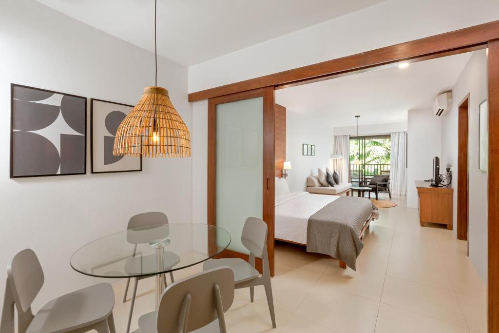 Mūsdienīga viesnīcas numura iekārtojums ar stikla galdu, ērtām krēsliem un gaisīgu interjeru. Logi sniedz skatu uz dārzu, radot relaksējošu atmosfēru.