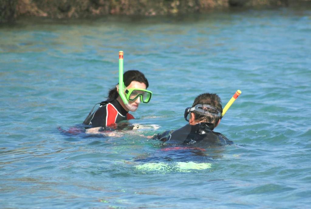 Divi cilvēki snorkelē šajā skaidrajā ūdenī, valkājot peldēšanas aprīkojumu un maskas. Viņi izskatas priecīgi un izpēta ūdens pasauli.