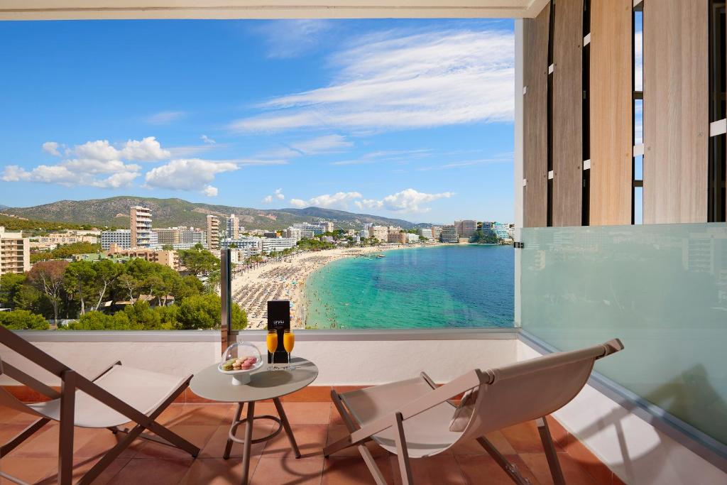 Skats no viesnīcas balkona uz pludmali un jūru, ar saulainu debesi un kalniem fona. Uz galda ir dzērieni un uzkodas, radot relaksējošu atmosfēru.