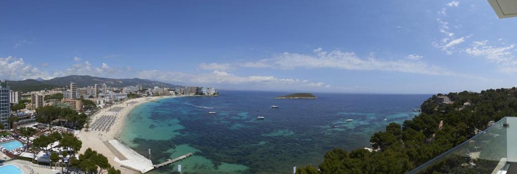 Krāšņa pludmales ainava ar skatu uz zilo jūru un apkārtējo piekrasti, kur redzami pludmales krēsli un viesnīcas. Debesis ir skaidras un saulainas, pievēršot uzmanību skaistajai dabai.
