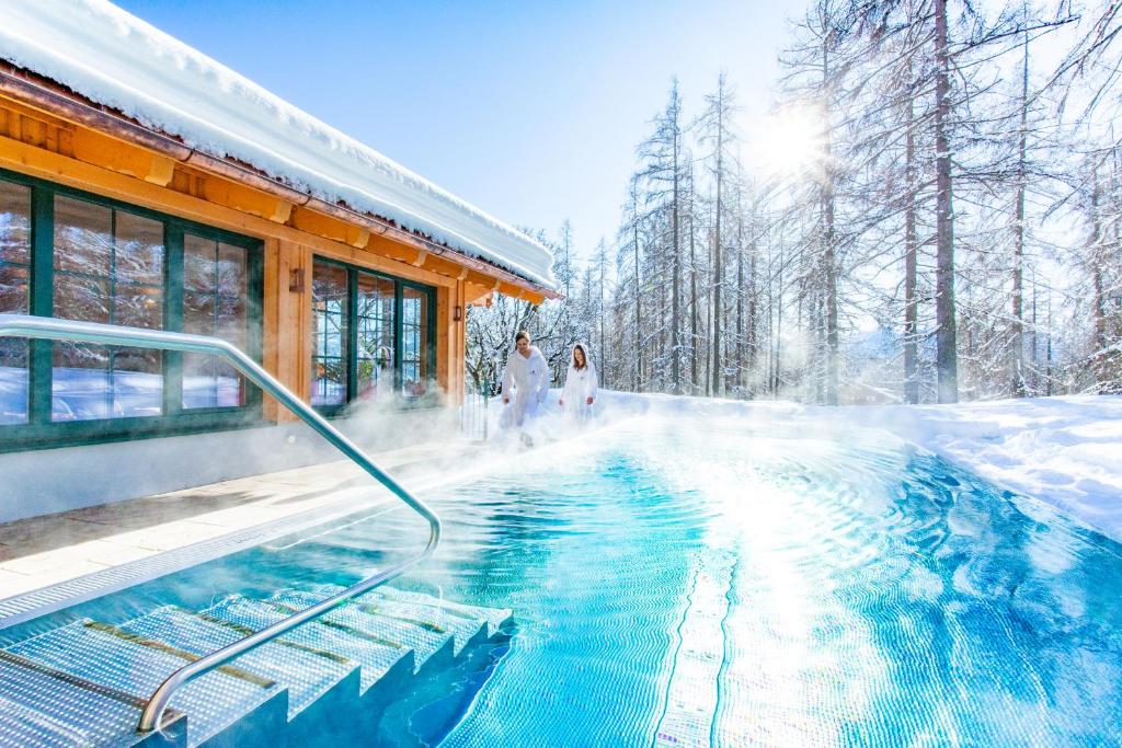 Slēpošanas kūrorts ar siltu baseinu sniegotā ainavā, kur divi cilvēki relaksējas, valkājot baltas drēbes. Sniega klāta apkārtne un saulainā debesis rada mierīgu atmosfēru.