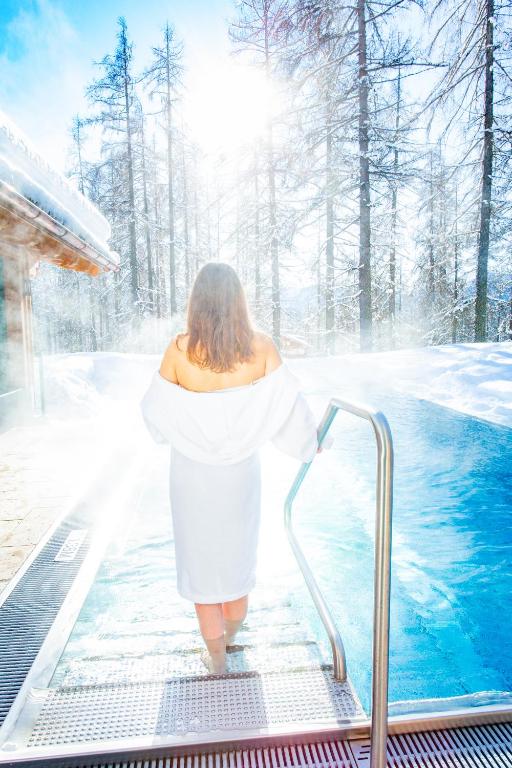Sniegots skats uz sievieti, kas ieiet karstā baseinā ziemas ainavā, apņemta ar tvaiku un saules gaismu. Aiz viņas redzami sniegoti koki un skaidrā debesis.