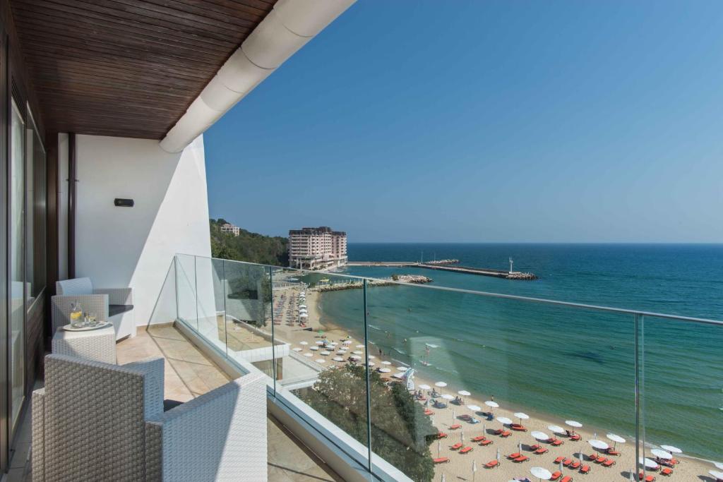 Skats no balkona uz pludmali ar saulainu debesi un mierīgu jūru. Redzami pludmales sauļošanās krēsli un viesnīcas komplekss.
