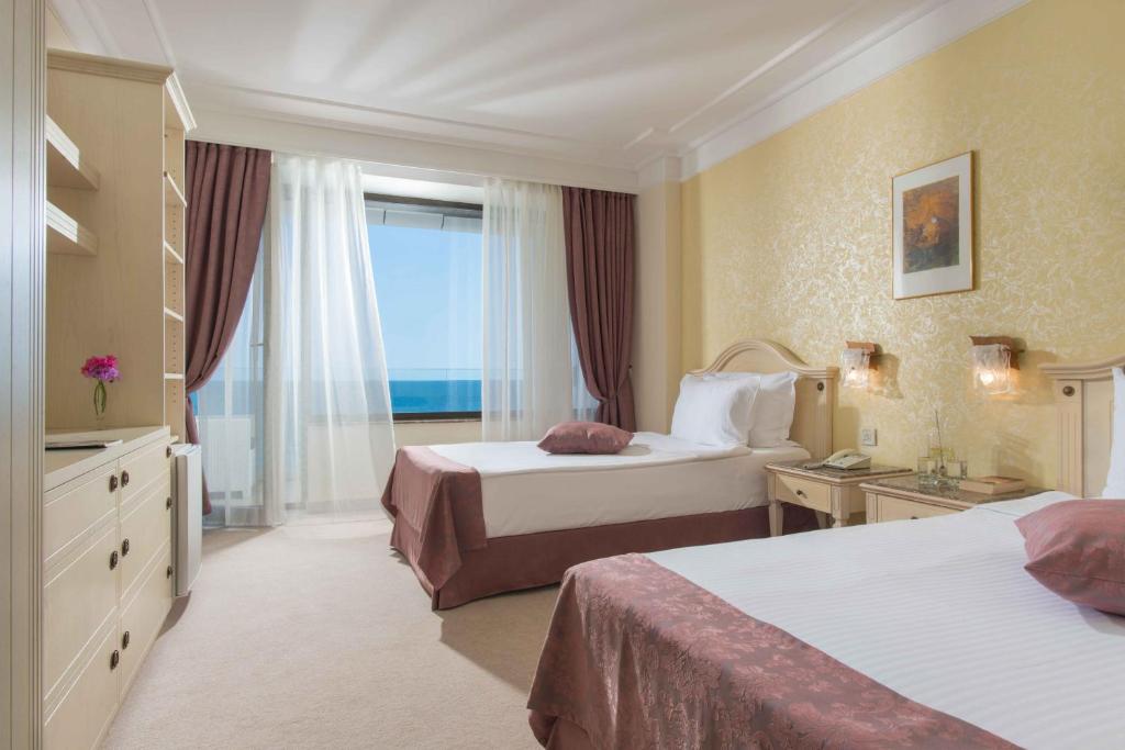 Gaiša viesnīcas numura iekārtojums ar divām gultām un skatu uz jūru. Telpā ir eleganta dekorācija un lielas logu aizkaru.