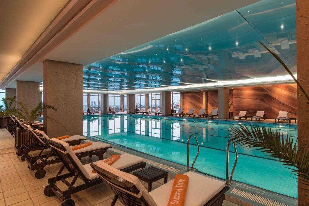 Iekštelpu peldbaseins ar modernu dizainu, kas iekļauj atpūtas krēslus un lielas logus, kas piedāvā skatu uz pilsētu. Telpas apgaismojums un zilā krāsa rada relaksējošu atmosfēru.