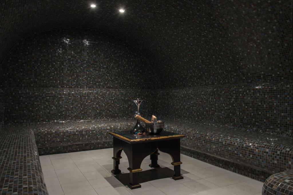 Mūsdienīgs tvaika pirts interjers ar melnas mozaīkas flīzēm un komfortablām sēdvietām. Uz galda atrodas šampanieša pudele un glāzes, radot relaksējošu atmosfēru.