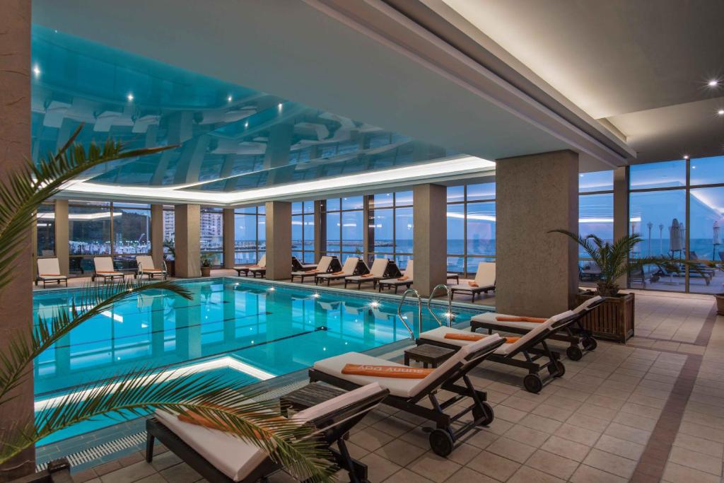 Iekštelpu peldbaseins ar modernu dizainu, apkārt peldbaseinam atrodas sauļošanās krēsli un lieli logi, kas sniedz skatu uz ārējo ainavu.