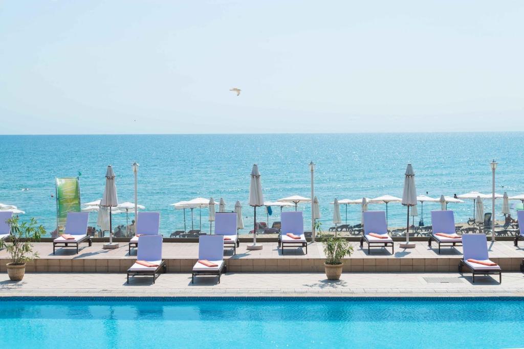 Pludmales atpūtas zona ar sauļošanās krēsliem un saulessargiem, skats uz mierīgu jūru un skaidru debesu. Idealizēta vasaras atmosfēra.