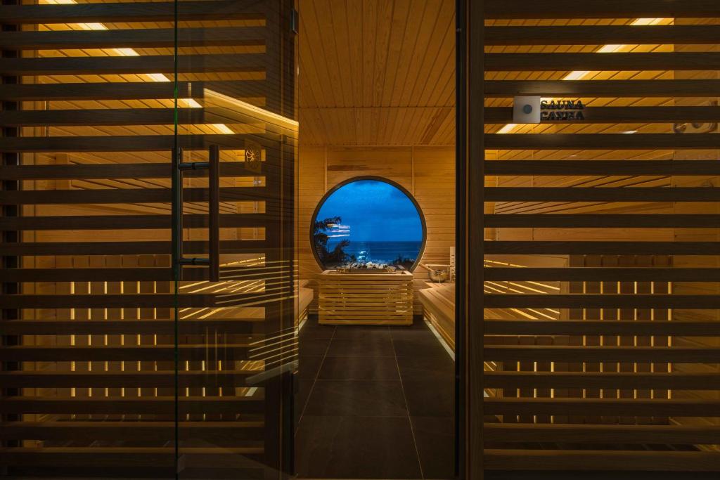 Mūsdienīga sauna ar koka interjeru un lielu apaļu logu, caur kuru redzams jūras skats. Siltas gaismas akcenti rada relaksējošu atmosfēru.