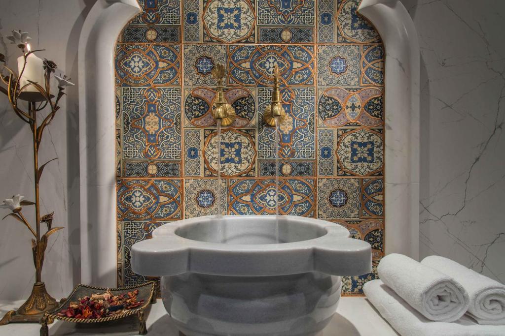 Elegants marmora baseins ar zeltītām krāna svirām, apņemts ar krāšņu flīžu sienu. Blakus atrodas dekoratīvs svečturis un salocītas dvieļi, radot relaksējošu atmosfēru.