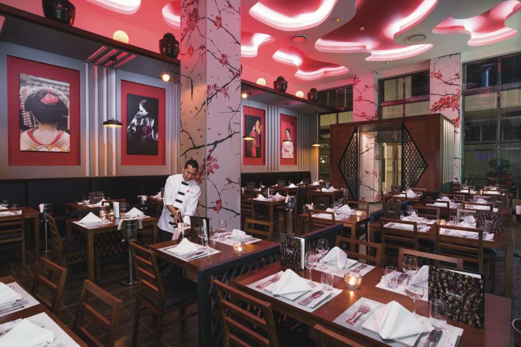 Mūsdienīgs restorāns ar elegantu interjeru, kurā ir sarkanas apgaismojuma detaļas un mākslinieciskas fotogrāfijas uz sienām. Galdi ir sakārtoti ar baltām salvetēm un glāzēm, radot izsmalcinātu atmosfēru.