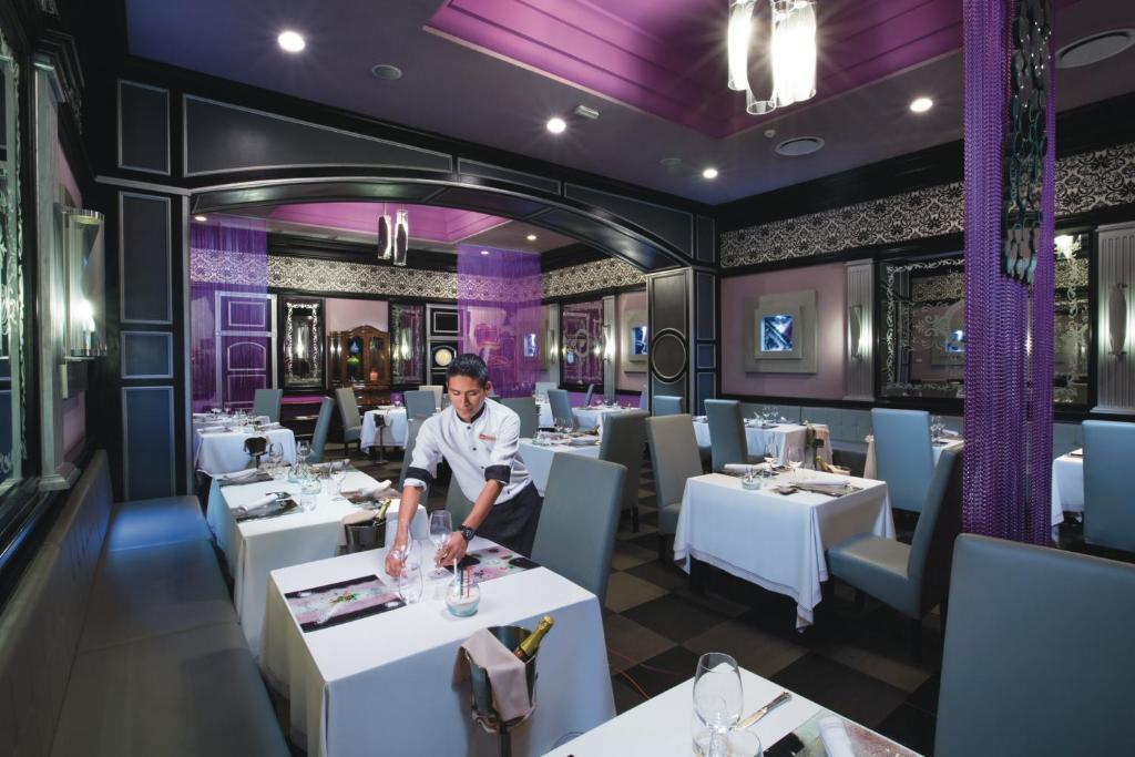 Elegants restorāns ar modernu interjeru, kurā apkalpojošais personāls sagatavo galdu. Interjers ir veidots ar purpura un melnām krāsām, radot stilīgu atmosfēru.