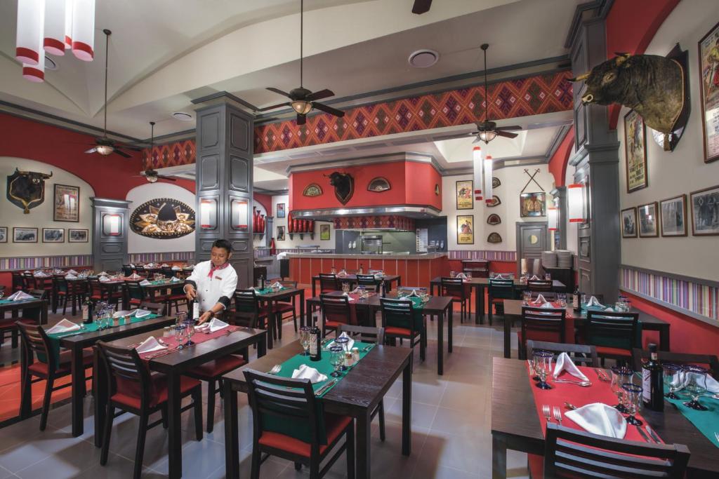 Mūsdienīgs restorāns ar sarkanu un pelēku interjeru, kurā ir koka galdi un krēsli, ar galdu klājumu un dekoratīvām fotogrāfijām uz sienām. Restorāna darbinieks sagatavo galdu apkalpošanai.