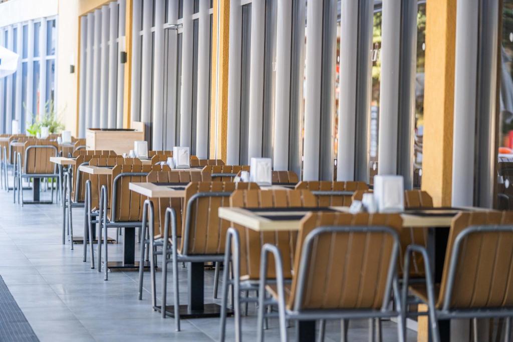 Āra kafejnīcas terase ar koka galdiem un krēsliem, kas gaida apmeklētājus. Rūpīgi sakārtoti galdi un maza zaļa auga dekorācija.