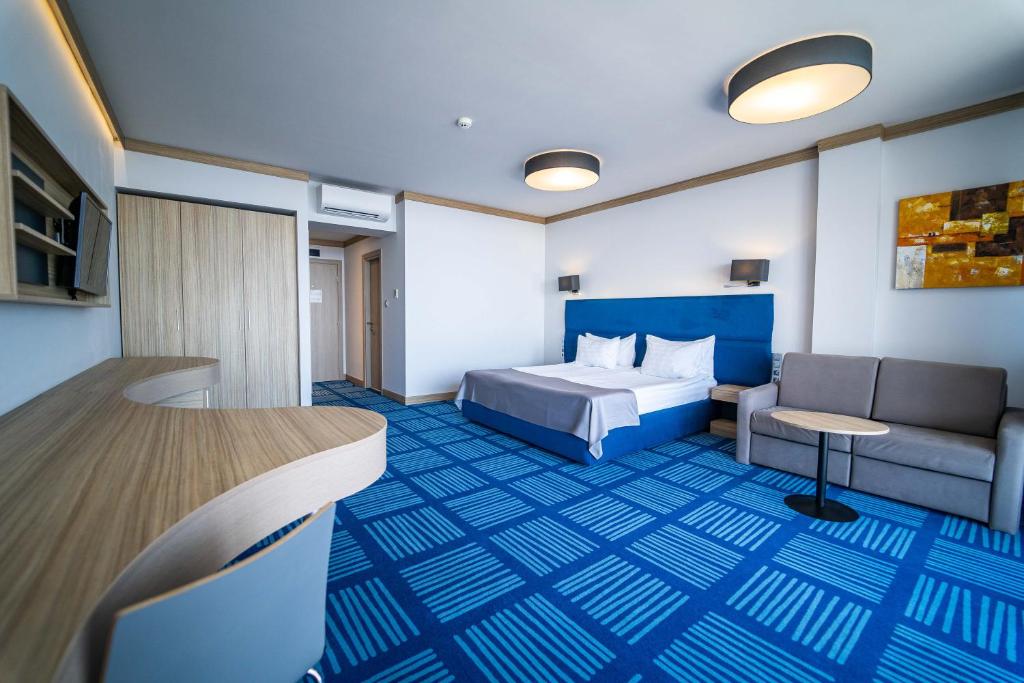 Mūsdienīgs viesnīcas numurs ar divguļamo gultu, dīvānu un rakstāmgaldu. Interjers ir gaišs un stilīgs, ar zilu paklāju un modernām lampām.