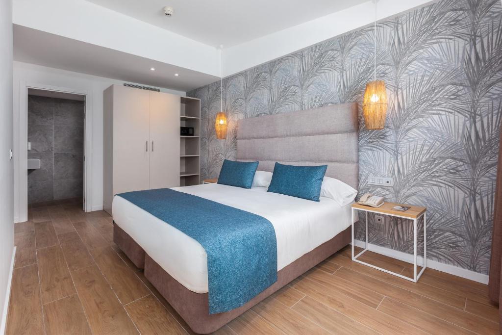 Mūsdienīga guļamistaba ar elegantu dizainu, kurā ir liela gulta ar zilu gultas pārklāju un dekoratīvām spilveniem. Sienas rotā stilizēts augu motīvs, un telpā ir dabīga koka grīda.