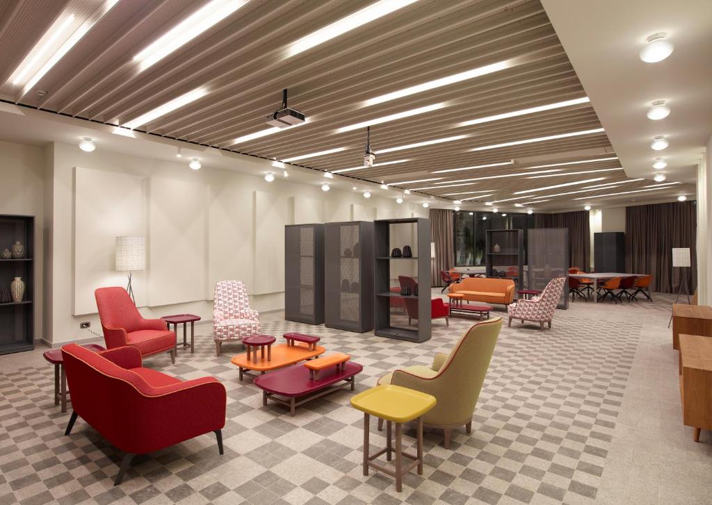 Mūsdienīgs koplietošanas telpas interjers ar krāsainiem krēsliem un galdiem, kas rada patīkamu un viesmīlīgu atmosfēru. Telpa ir apgaismota ar modernām lampām un ir aprīkota ar stilīgām mēbelēm.