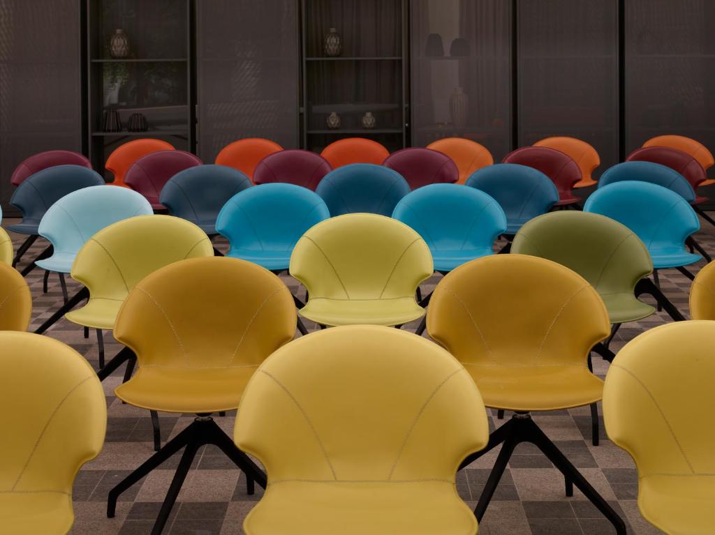 Krāsainu krēslu rinda modernā telpā, kas izceļ spilgtas dzeltenas, zilas, sarkanas un oranžas krāsas. Radošs un stilīgs interjera elements.