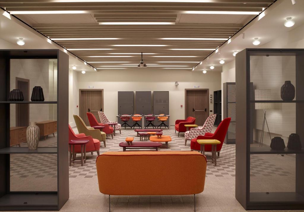 Mūsdienīgs atpūtas telpas interjers ar krāsainām mēbelēm un stilīgām dekorācijām. Telpā ir sarkanas, oranžas un dzeltenas krēsli, kā arī elegantas vitrīnas.