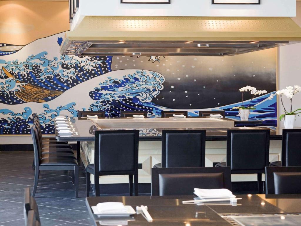 Modernas japāņu restorāna interjers ar elegantām melnām krēslām un košām viļņu gleznām uz sienas, radot relaksējošu atmosfēru. Uz galda sakārtotas ēdienu pasniegšanas piederumi.