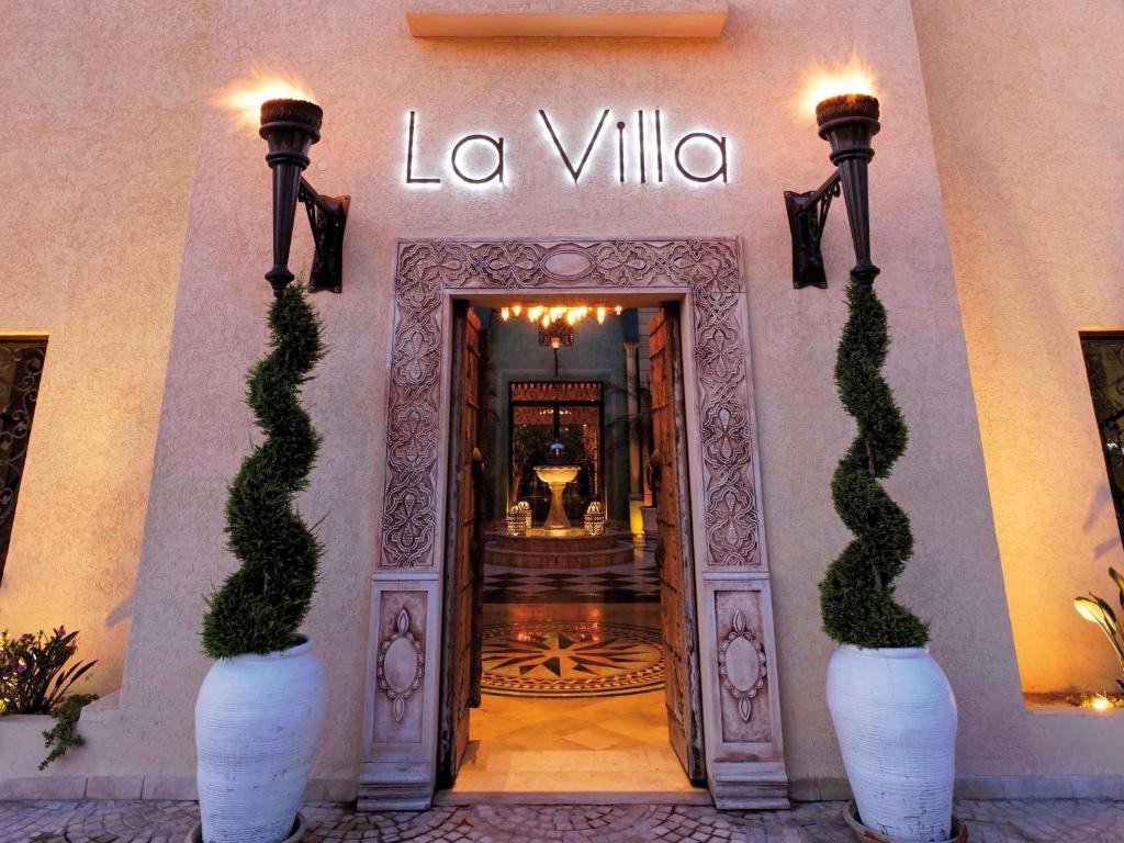 Elegantā ieeja restorānā "La Villa" ar greznu apgaismojumu un dekoratīvām puķu podiem, kas izceļ modernu arhitektūru un viesmīlīgu atmosfēru.