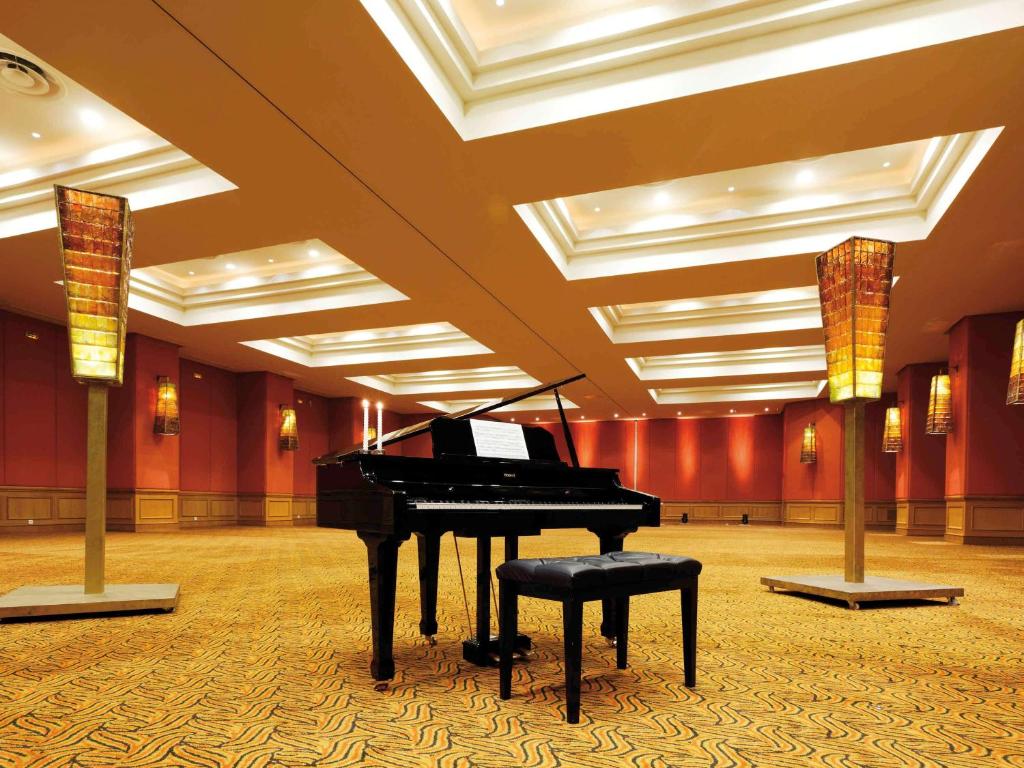 Melodiska klavieres stāv tukšā telpā ar siltu apgaismojumu un stilīgām lampām, radot elegantu atmosfēru. Telpas interjers ir moderns ar bagātīgu krāsu paleti.