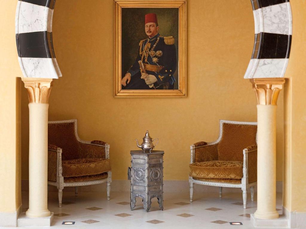 Eleganta viesistaba ar diviem krēsliem un metāla galdiņu, pie sienas atrodas krāšņs portrets. Interjeram raksturīgas siltas krāsas un grezni dekoratīvi elementi.