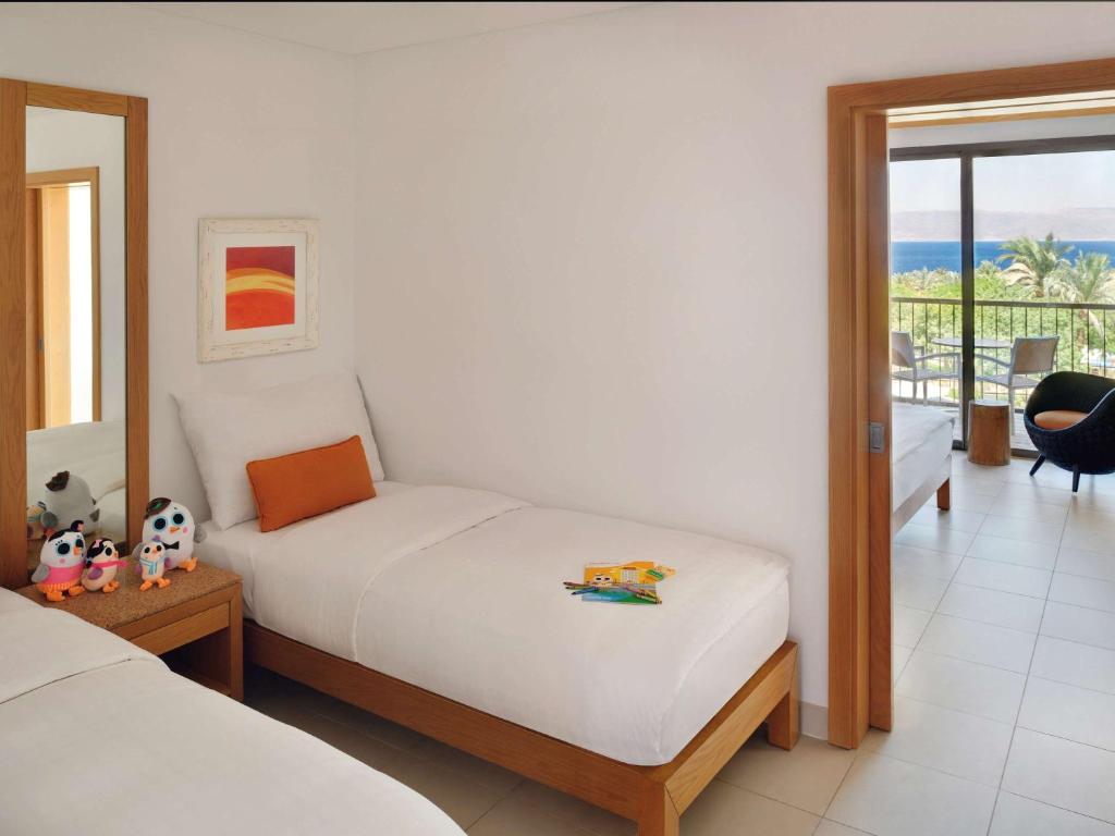 Mūsdienīga viesnīcas istaba ar divām gultām, spilvenu un spilvenu ar oranžu apdari. Uz galda atrodas mīļlietiņas, bet logu skatā redzama ainava ar jūru un zaļumiem.