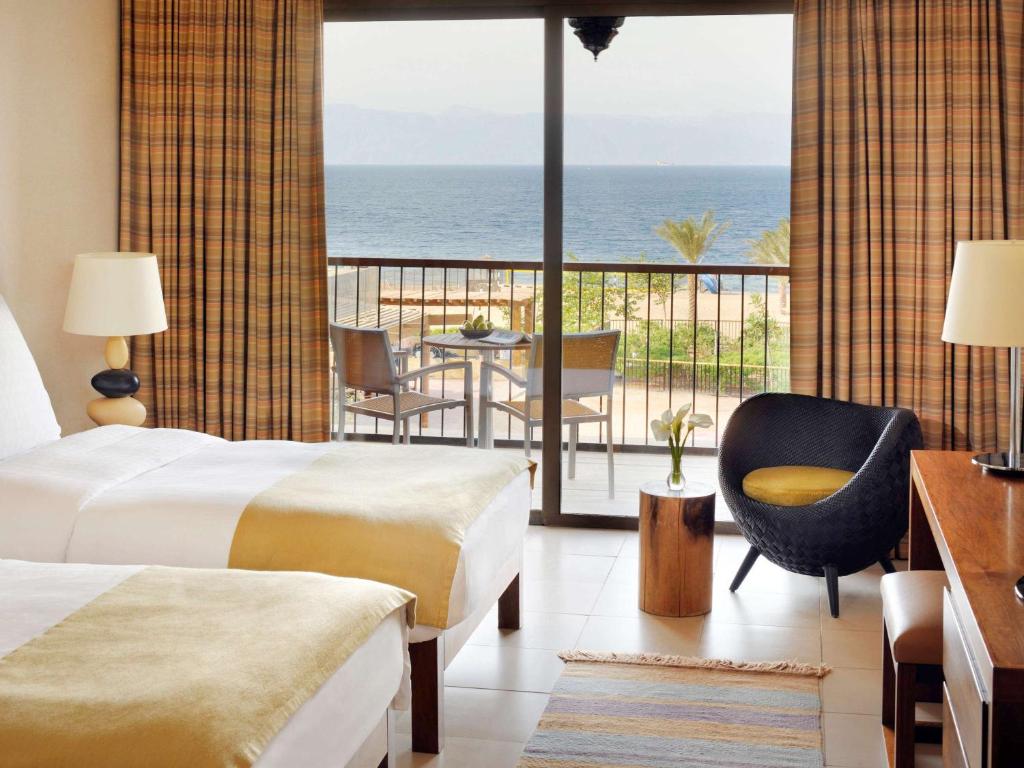 Mūsdienīgs viesnīcas numurs ar skatu uz jūru, divām gultām un ērtu sēdvietu. Logu aizkari un gaišas krāsas interjers rada patīkamu atmosfēru.