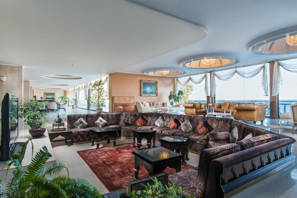 Elegants un plašs viesistabas interjers ar greznām mēbelēm un augiem. Telpā ir lieli logi, kas nodrošina dabisko apgaismojumu un skatu uz ārpusi.