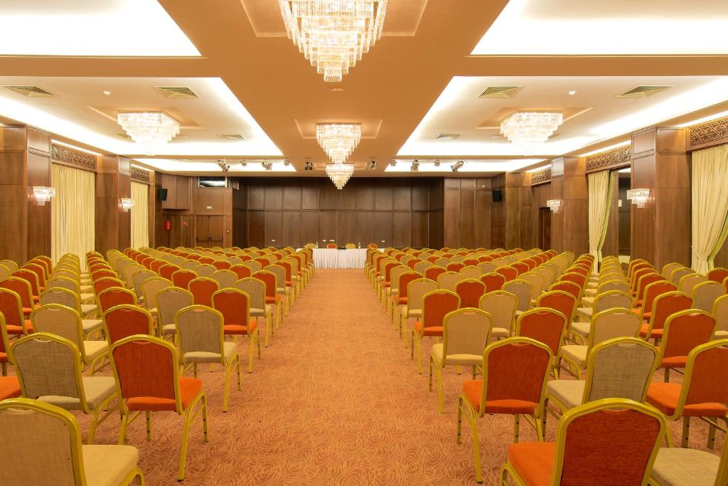 Konferenču zāle ar rindu sēdvietām un elegantiem lustriem, gatava pasākumam. Telpa ir plaša un labi apgaismota, ar koka apdari un komfortablām krēslu rindām.