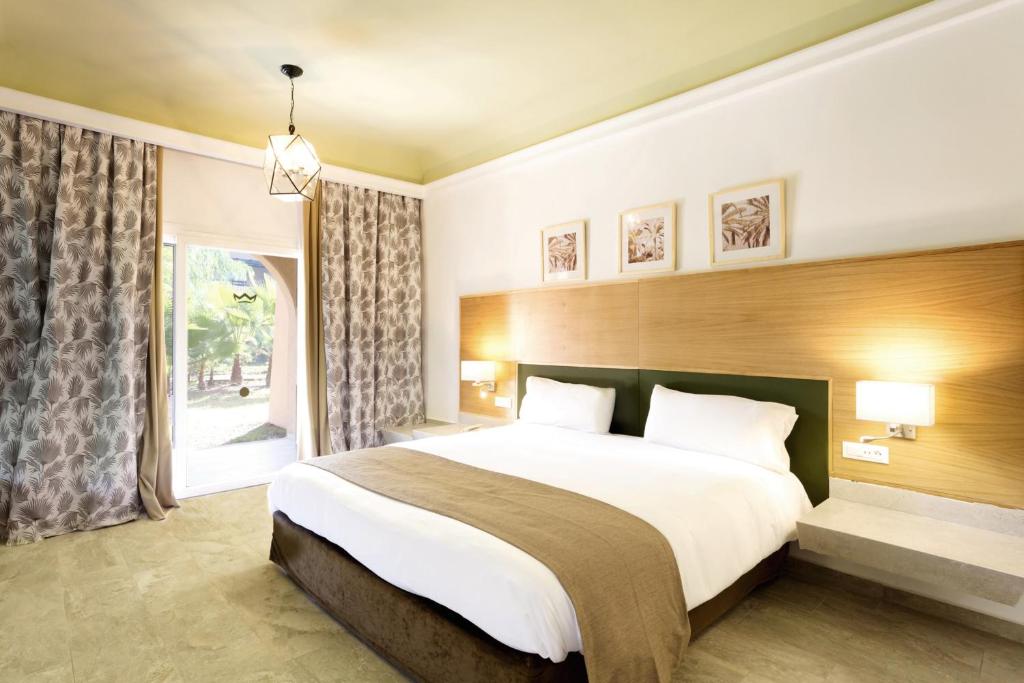 Mūsdienīgs guļamistabas interjers ar divguļamo gultu, dabīgām krāsām un gaišiem aksesuāriem. Logi ar aizkariem, kas ved uz dārzu, un mājīga apgaismojuma elementi papildina telpas atmosfēru.