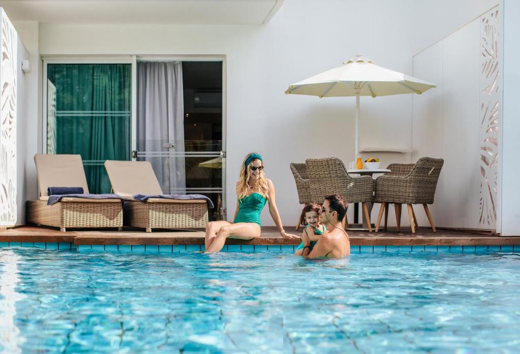 Ģimene bauda atpūtu pie baseina, māte sēž uz malas, tērpta zaļā peldkostīmā, bet tēvs tur bērnu ūdenī. Fons ietver ērtus sauļošanās krēslus un ēdamgaldu zem lietussarga.