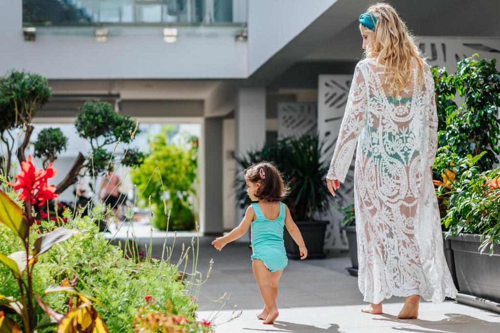 Māte un mazulis pastaigājas pa saulainu dārzu, ko ieskauj krāsaini ziedi un zaļumi. Māte valkā baltu garu kleitu, bet mazulis ir tērpies gaišā peldkostīmā.