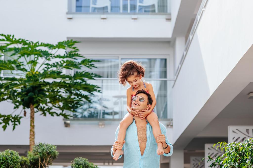 Tēvs ar meitu, kas sēž viņam uz pleciem, priecīgi pozē modernā ēkā ar zaļiem augiem fonā.