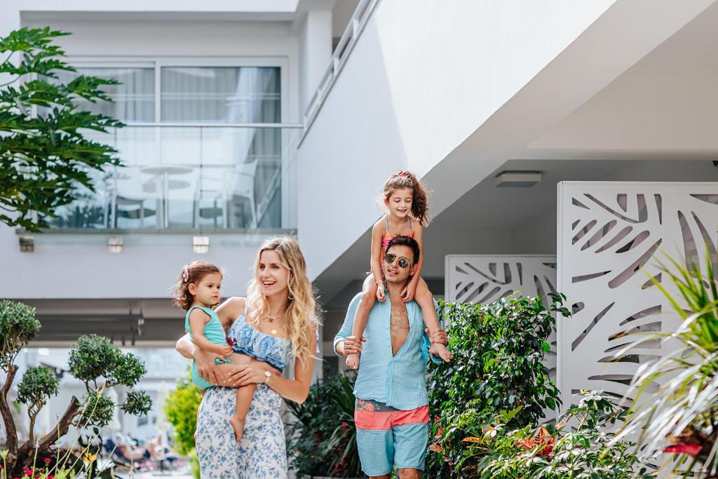 Ģimene izbauda laiku kopā, ejot pa saulainu viesnīcas teritoriju. Māte nes mazo meitu, bet tēvs ar smaidu nes otro meitu uz pleciem.