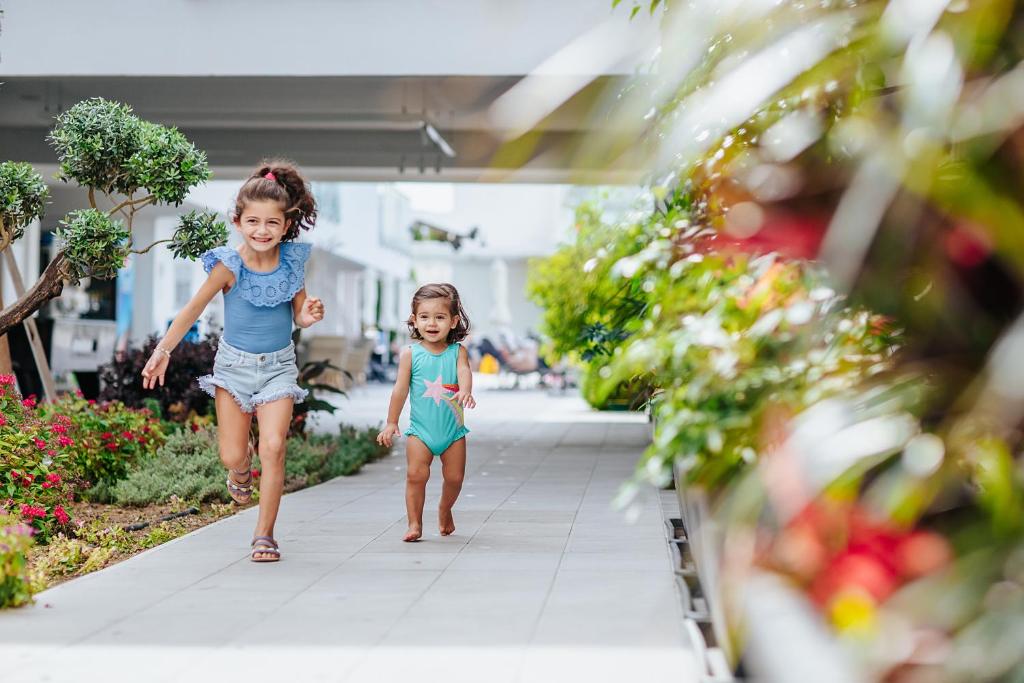 Divas meitenes priecīgi skrien pa ziedu rotātu celiņu, baudot saulainu dienu. Apkārt ir zaļi augi un ziedi, radot dzīvīgu un jautru atmosfēru.