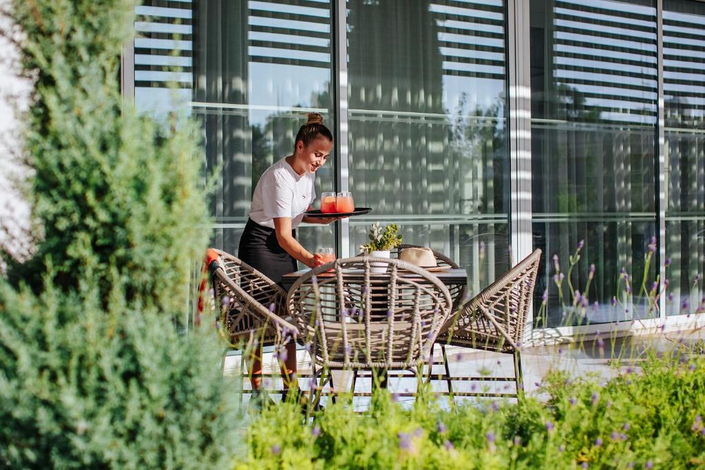Viesmīle pasniedz dzērienus āra terasē, kur blakus ir elegantas vītņu krēsli un zaļumi. Daba un moderns dizains rada patīkamu atmosfēru.