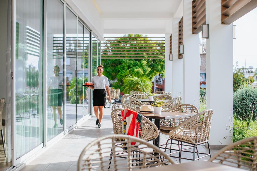 Viesmīle nes ēdienu uz terasi, kur ir moderni galdi un krēsli, kam apkārt zaļumi. Gaisīgs restorāns ar lieliem logiem un saulainu atmosfēru.