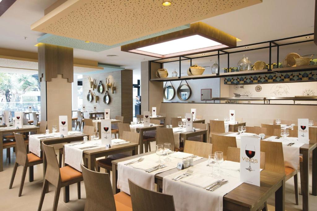 Moderna restorāna interjers ar elegantiem galdiem un krēsliem, uz kuriem ir izvēlnes. Uz galdiem ir glāzes un galda piederumi, radot viesmīlīgu atmosfēru.